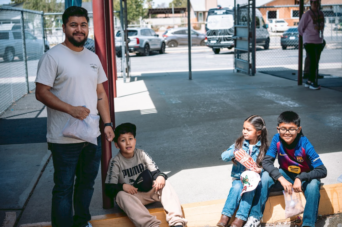Four children sitting on a curb in a parking lot with a man standing nearby.