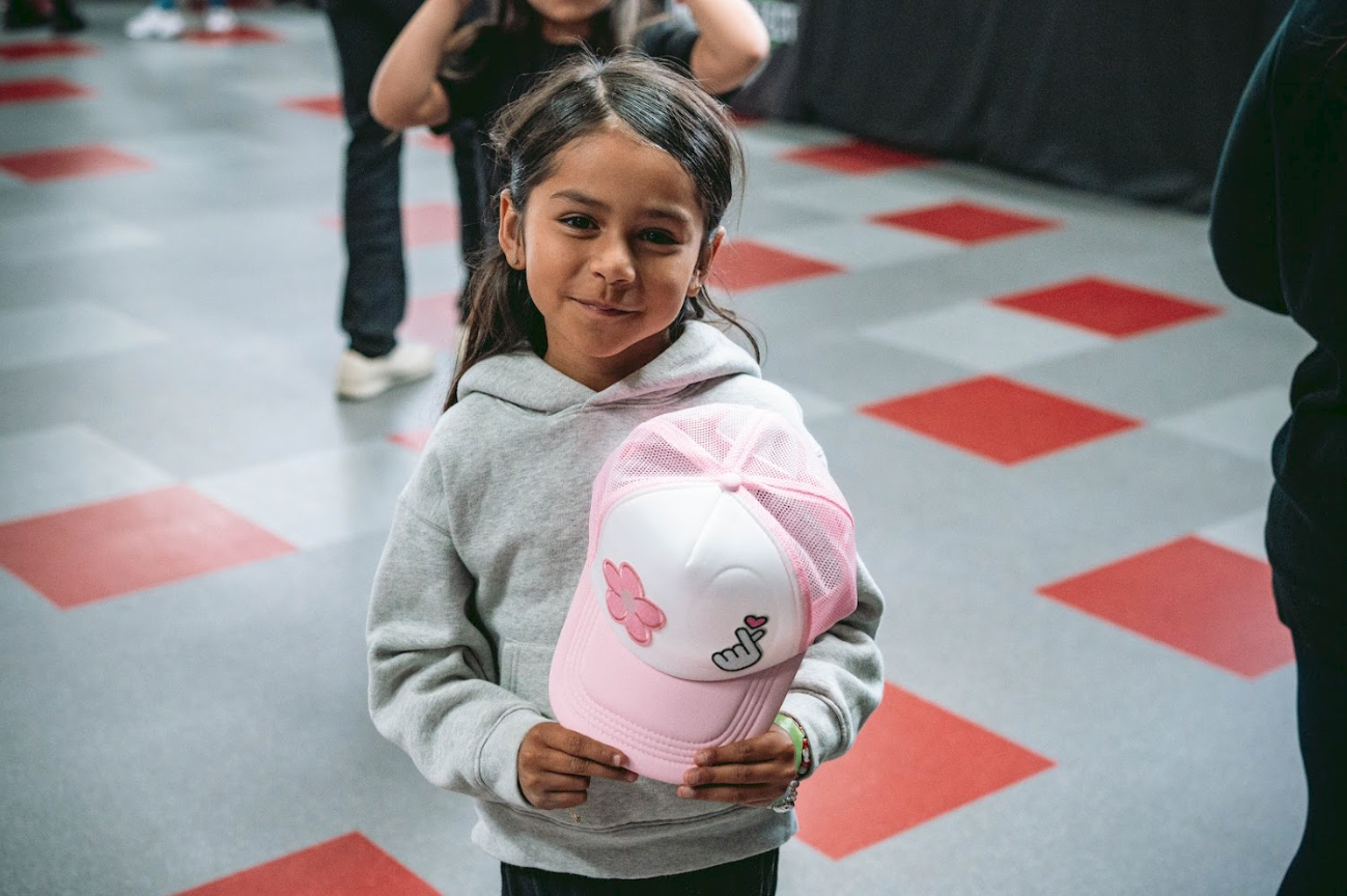 A young girl holding a pink and white cap with a butterfly and middle finger graphic, standing on a checkered floor, smiling at the camera.