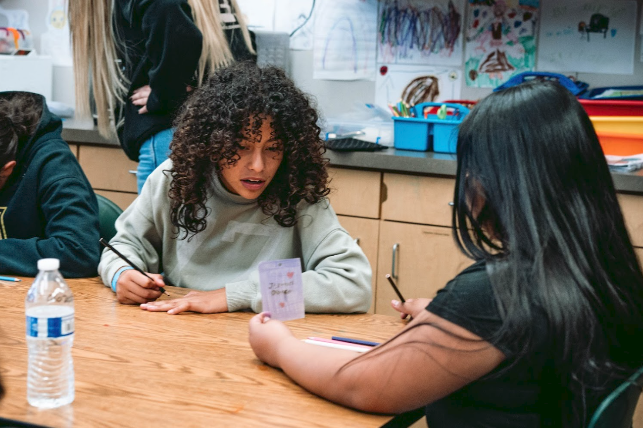 Two girls are sitting at a wooden table in a classroom, engaged in a conversation. One girl with curly hair and a beige sweater looks surprised or curious, while the other girl with straight black hair and glasses wears a black shirt. There are books