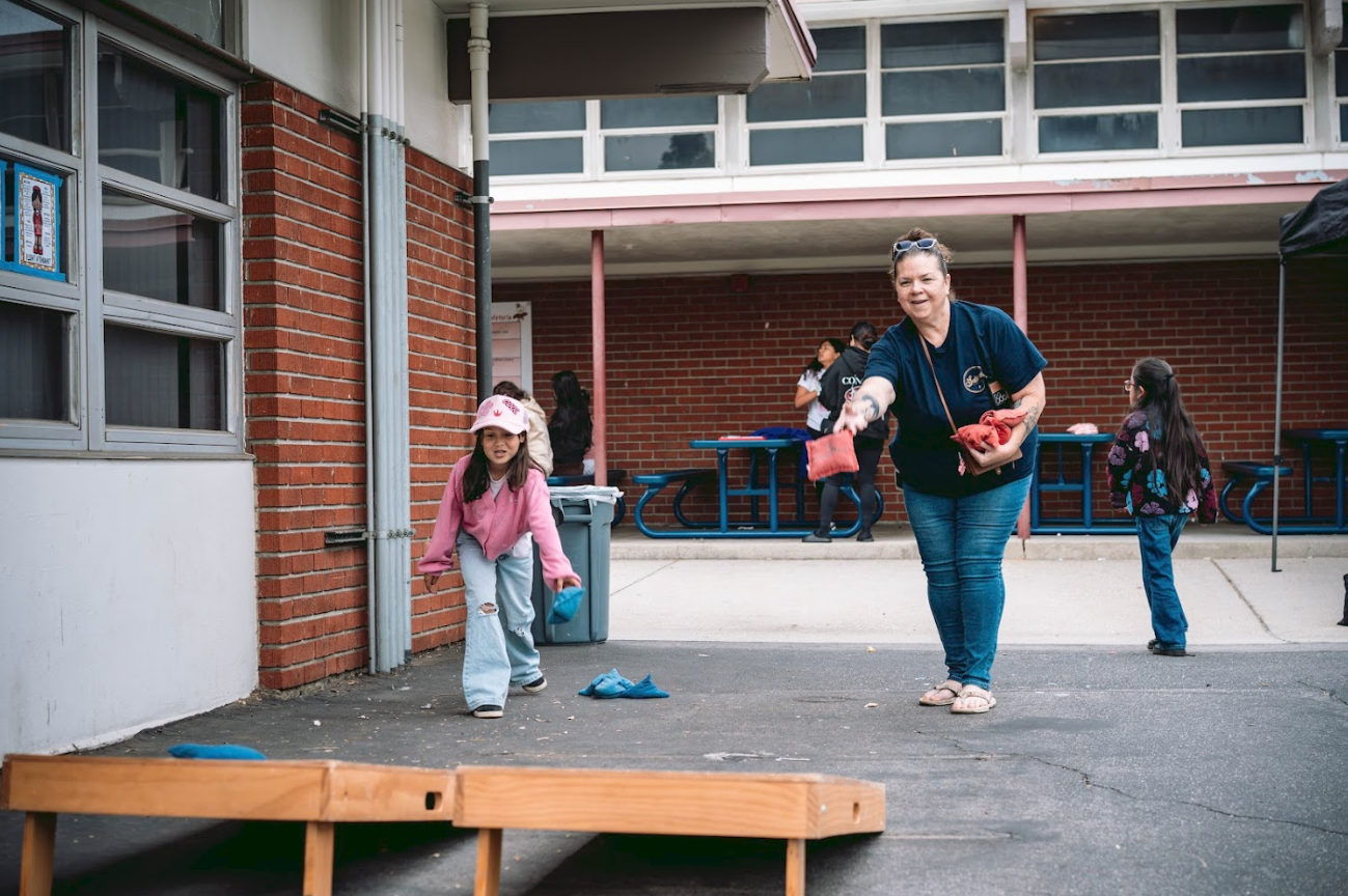 A woman and a young girl playing the game of cornhole outside a school building. The woman is reaching to toss a bean bag, and the girl is watching while holding another bean bag.
