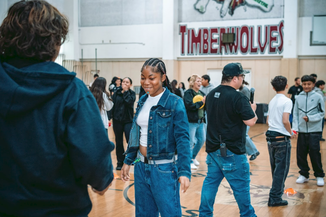 A girl in a denim jacket and jeans smiling at a game in a gymnasium with a Timberwolves sign on the wall and several people playing and talking.