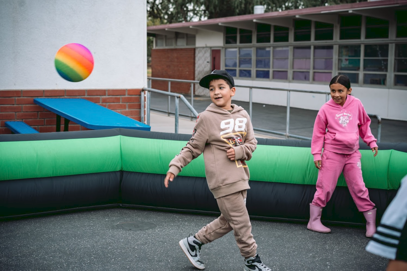 Two children, a boy and a girl, playing inside a bounce house outdoors near a school or community center. The boy is wearing a beige hoodie and matching sweatpants with a black cap and sneakers, while the girl is dressed in a pink hoodie and pink rai