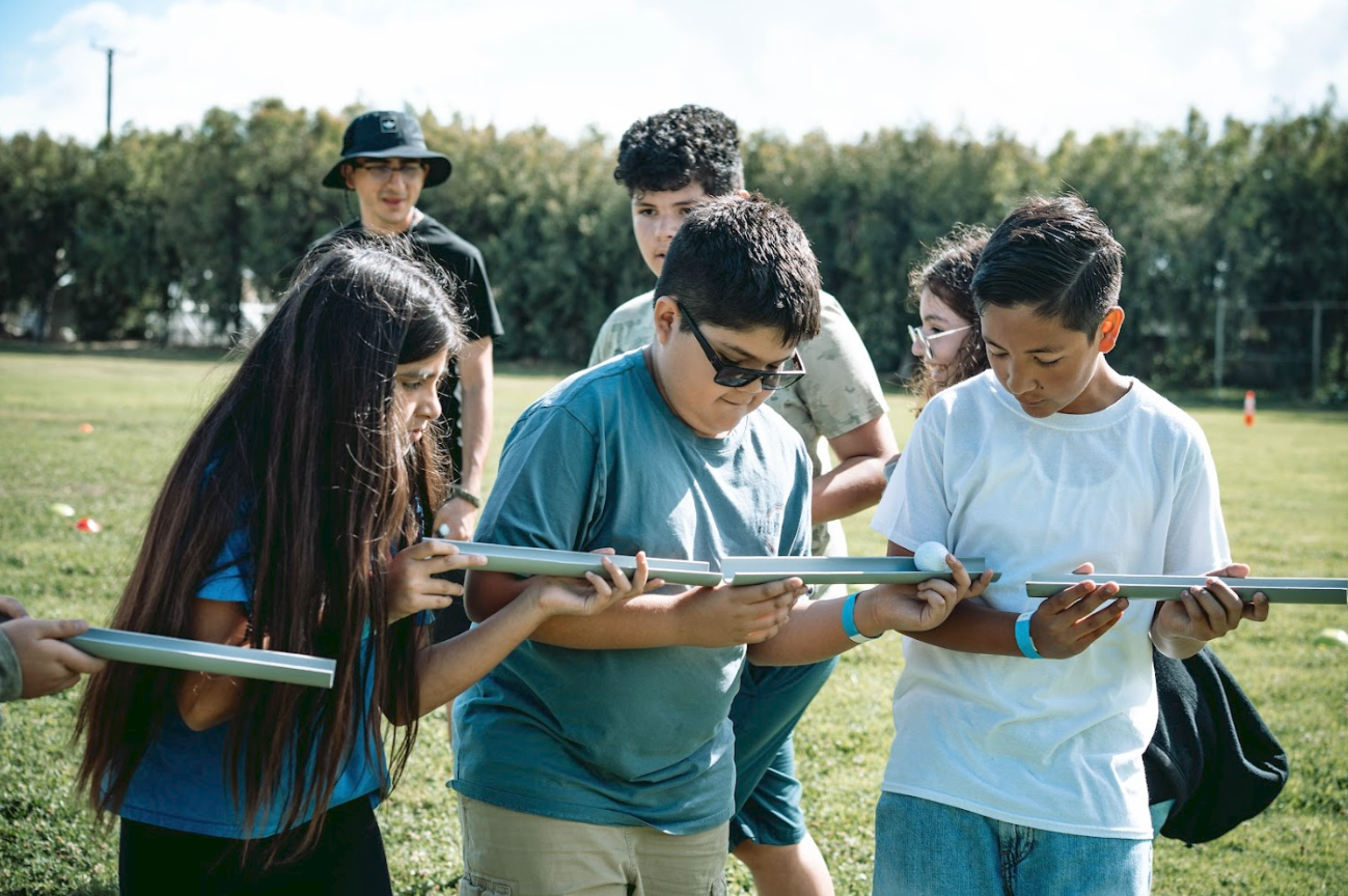 Group of children and a young adult outdoors examining a small white ball on a stick.