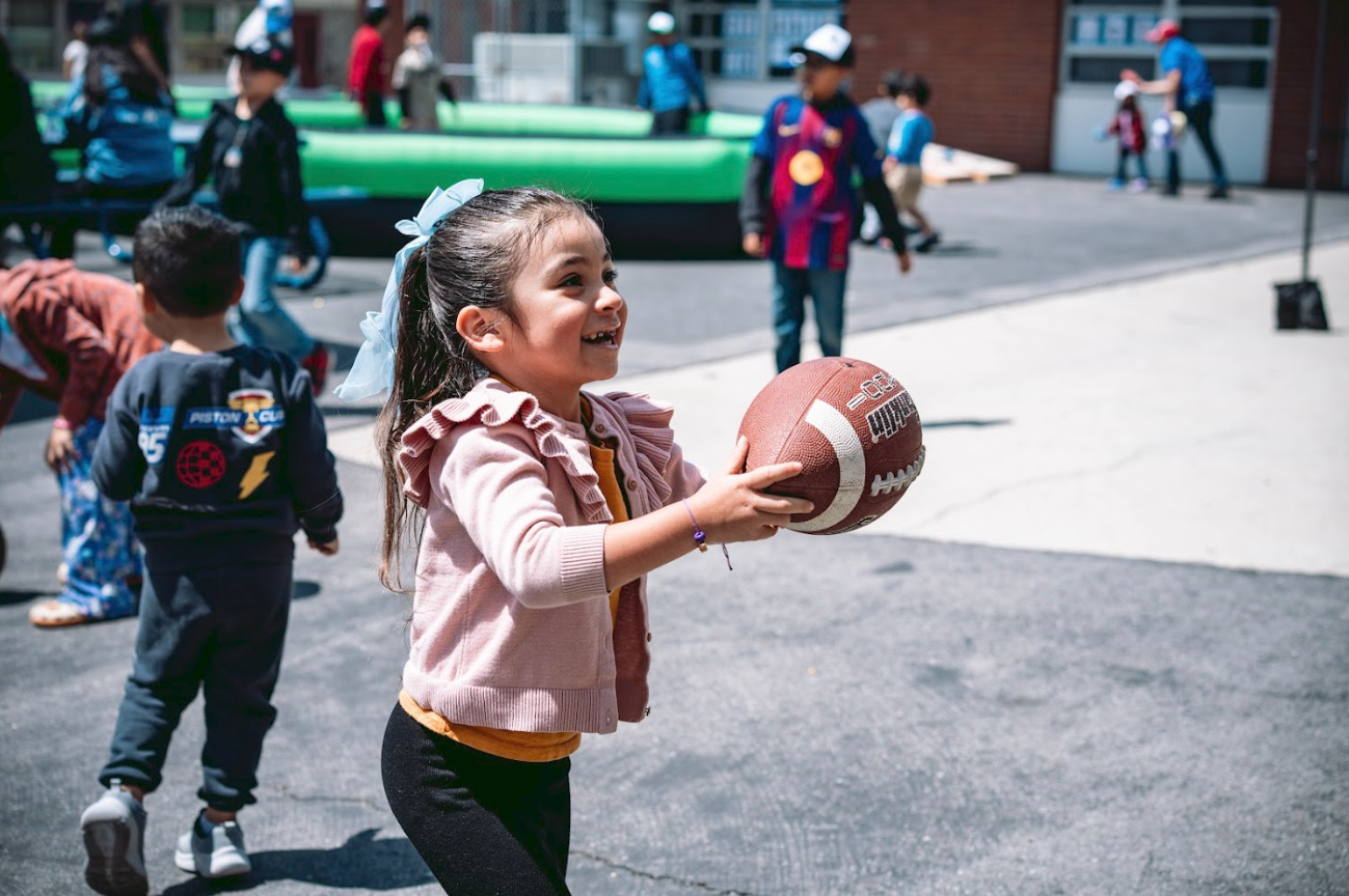 A young girl holding an American football, smiling, at a playground with other children playing in the background.
