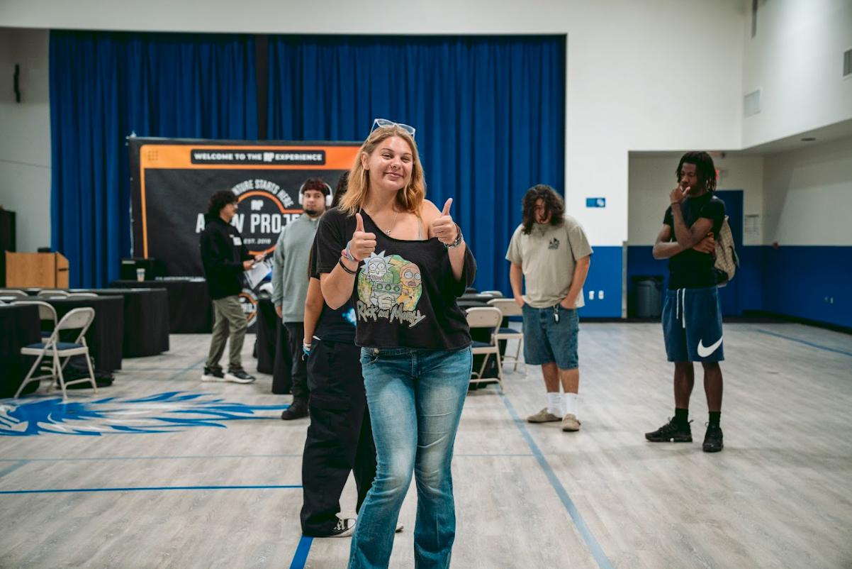 A group of young people in a gymnasium or event space, with a woman in the foreground giving a thumbs-up and smiling, and others standing in the background.