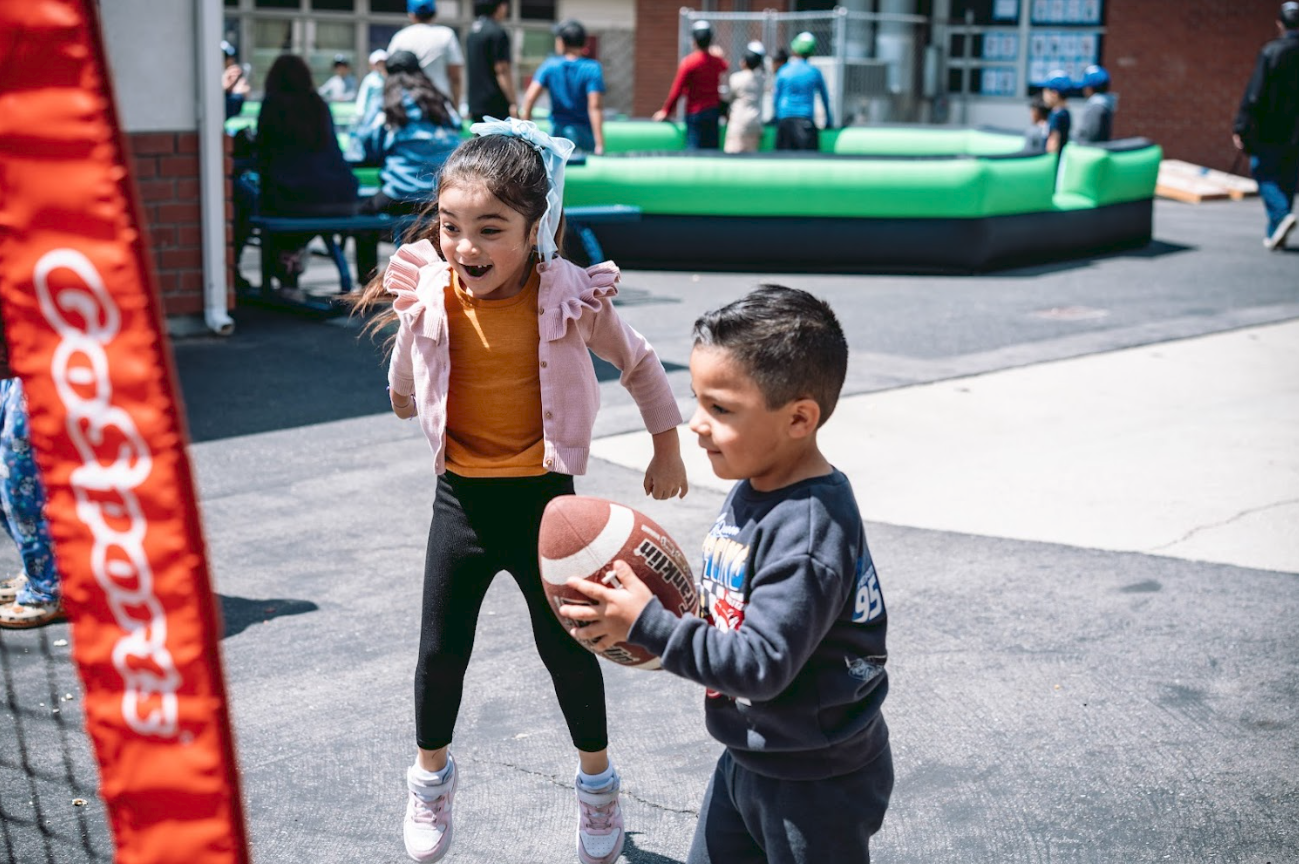 Kids playing basketball outdoors at a school or playground, with children in the background and a green inflatable structure.
