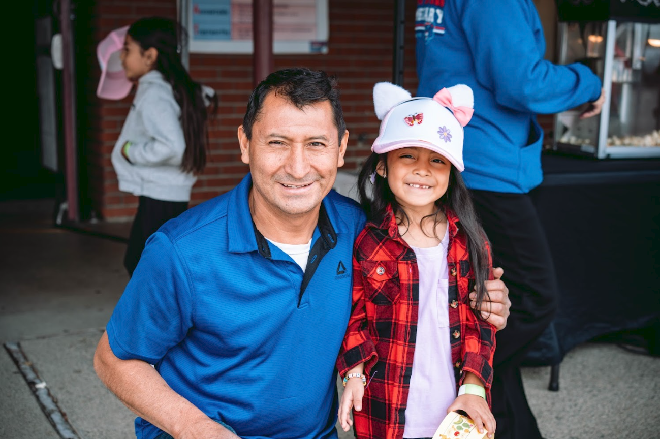 A man and a young girl smiling and posing together outdoors at a public event or fair. The man is kneeling with his arm around the girl, who is wearing a white hat with animal ears and colorful embellishments. In the background, a girl with a pink ca