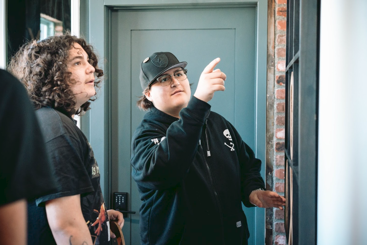 Two young men standing near a door, one pointing at something on a wall display, with the other looking on, inside a building with brick accents.