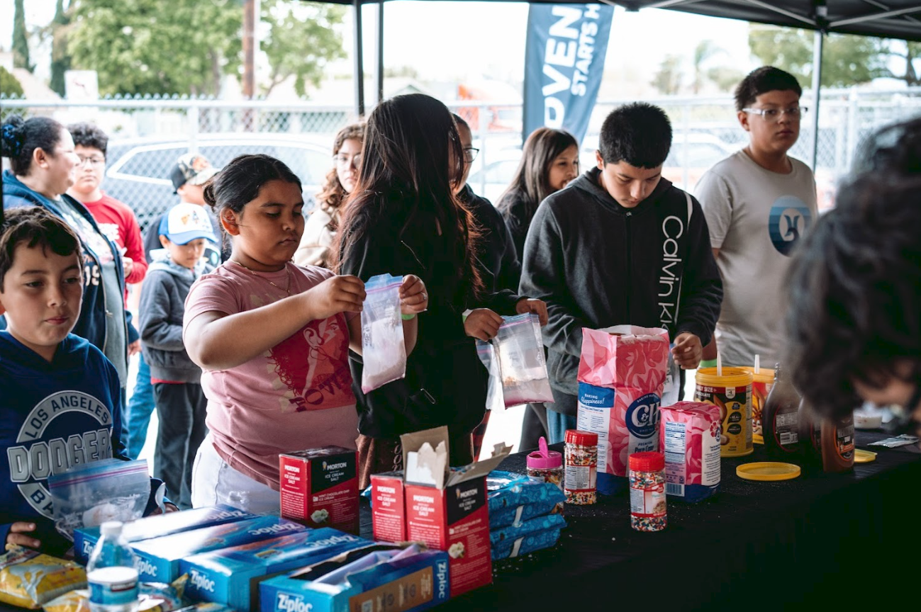 Group of children and adults gathered around a table under a canopy, dealing with baking supplies such as flour, sprinkles, and food coloring.