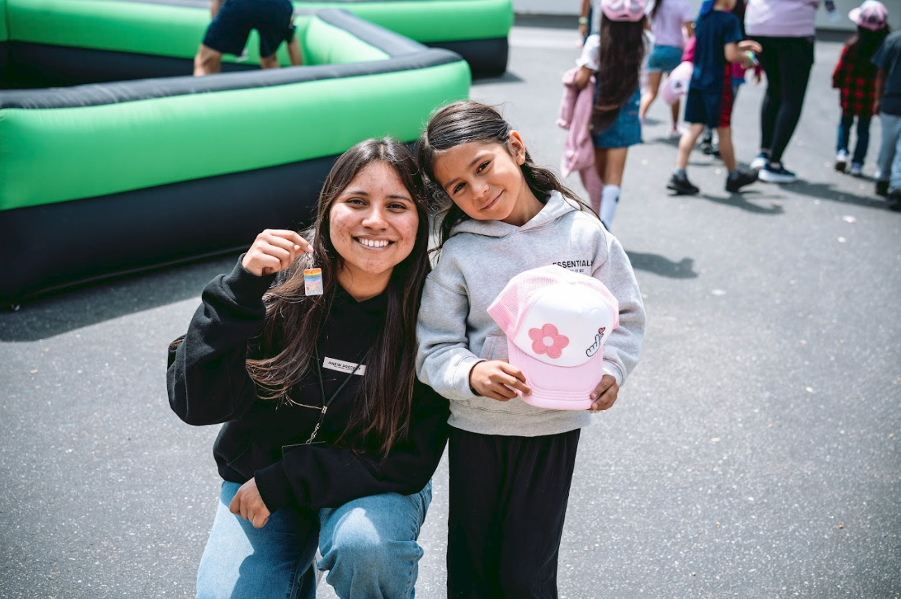 A young Hispanic girl and a young woman crouch together at an outdoor event, smiling at the camera. The girl holds a pink and white cap, and the woman holds a small rainbow sticker, with children and inflatable structures in the background.