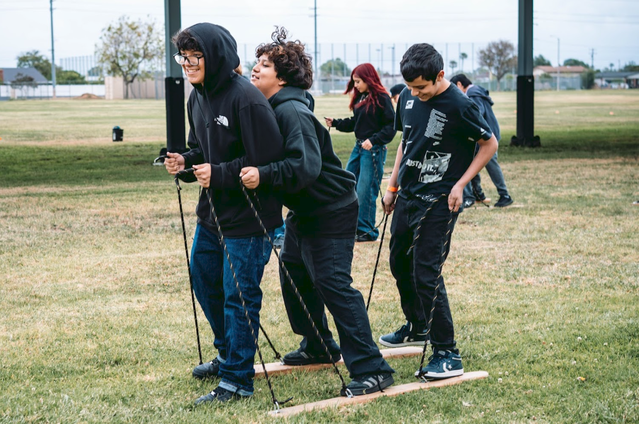 Group of children participating in outdoor team-building activity on grass field, standing on wooden planks connected with ropes, smiling and practicing coordination.