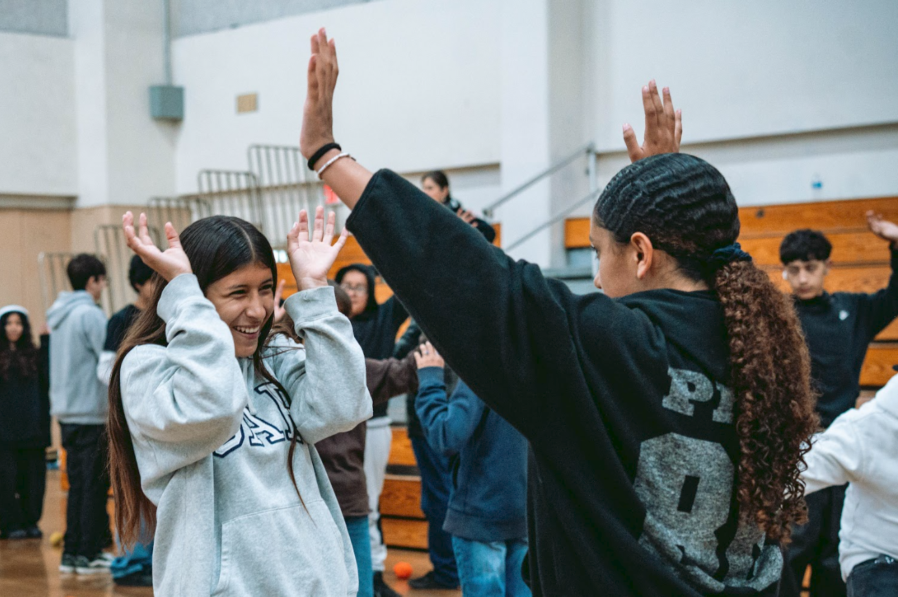 Two young girls in a gymnasium high-fiving each other, surrounded by other children.
