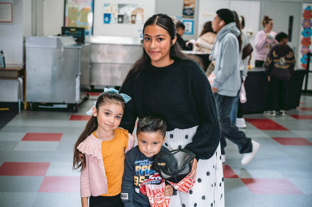 A woman with dark hair wearing a black sweater and a white skirt with black polka dots is standing in a school cafeteria with two children, a girl with long hair wearing a pink cardigan and orange shirt, and a boy with short hair wearing a blue sport