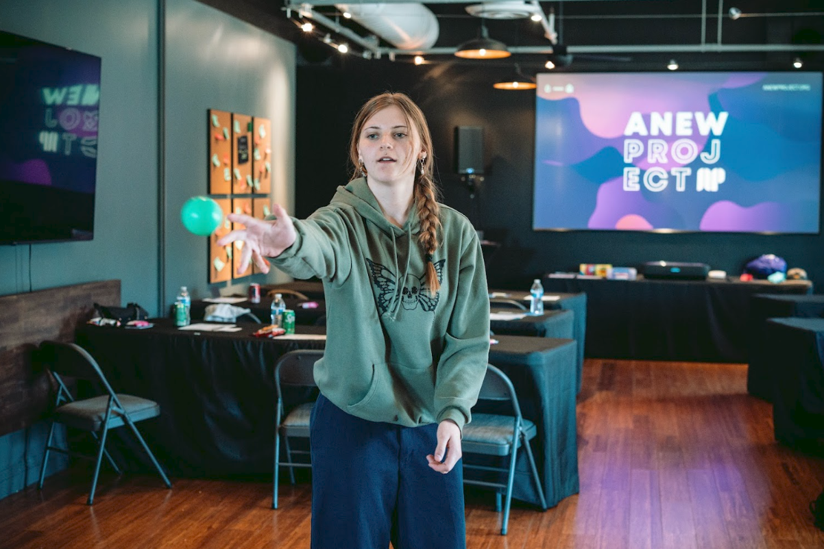 A young woman with long red hair in a braid, wearing a green hoodie with a butterfly design and blue jeans, is reaching out to catch a small green ball in a dimly lit room with tables, chairs, bottles of water, and a large screen displaying the words