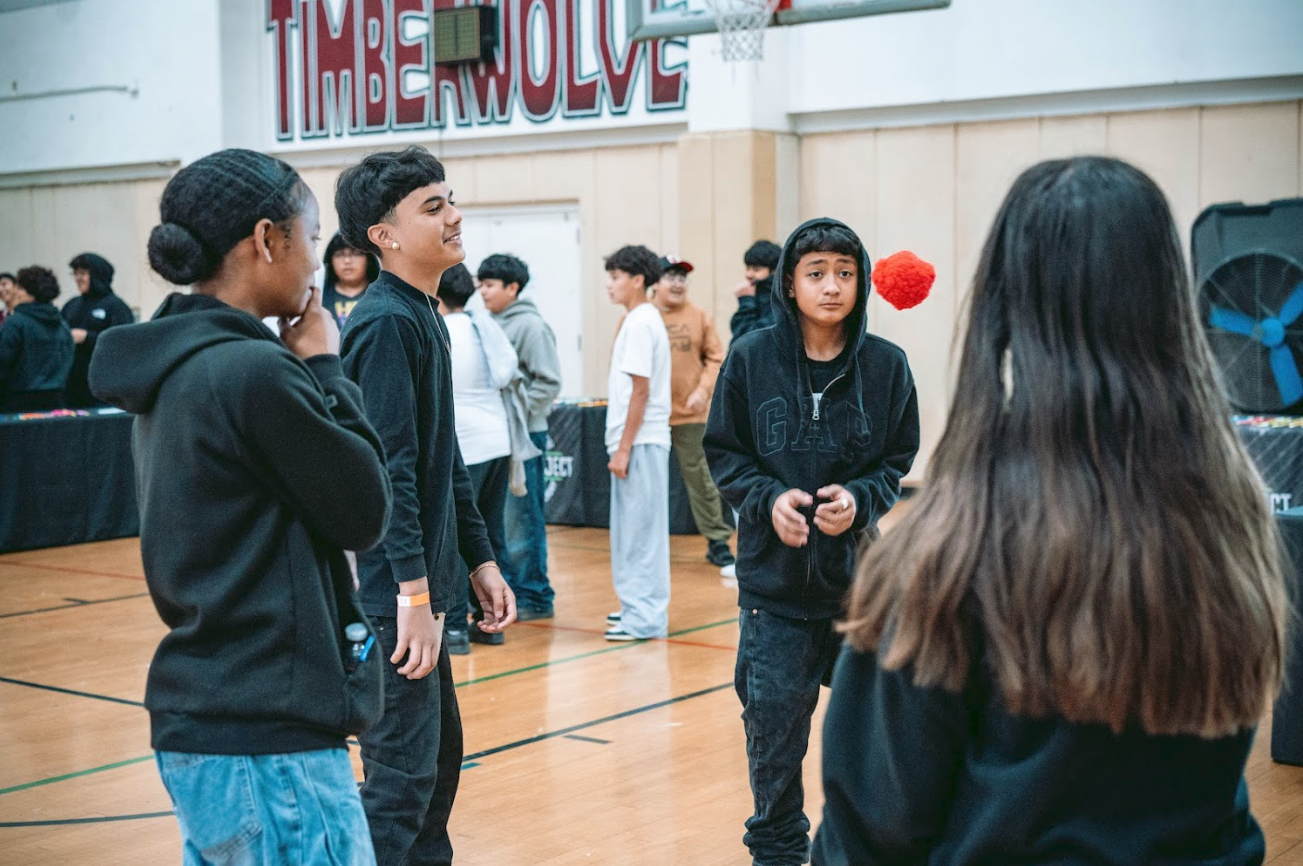 Group of diverse teenagers standing and talking in a gymnasium, with a red pom-pom flying through the air, and a banner reading 'TIMBERWOLVES' in the background.