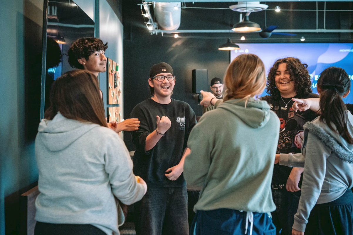 Group of diverse young people socializing and smiling in a modern indoor venue with dark walls, ceiling fans, and a large digital screen in the background.