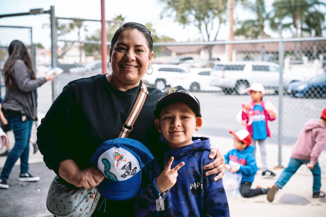 A woman and a young boy smiling at the camera outdoors, with children blurred in the background. The woman has dark hair tied back, wearing a black top, and holding a blue cap with a dinosaur and shark design. The boy is wearing a baseball cap, a nav