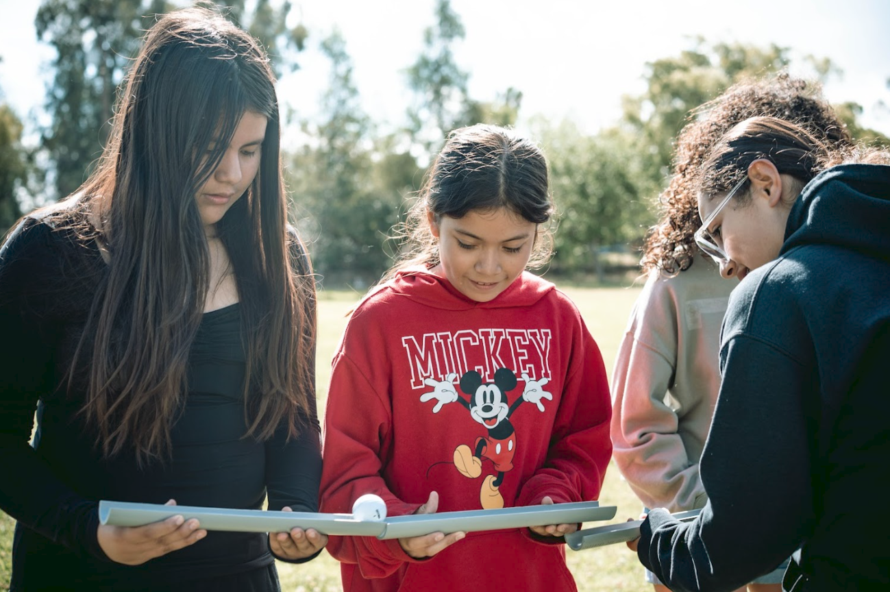 Four young girls standing outdoors on grass, looking at a white drone with propellers, in a park-like setting with trees in the background.