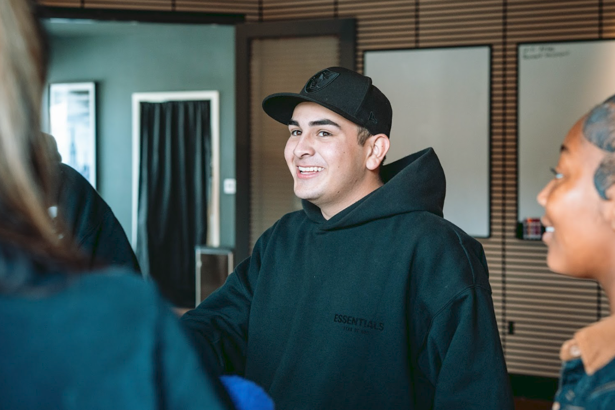 A young man wearing a black hoodie and a black cap smiling and talking to two women in an indoor setting.