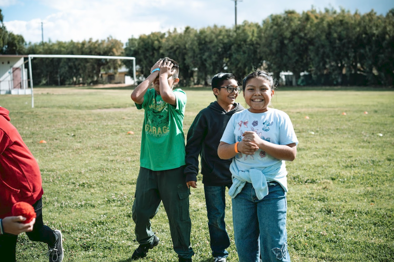 Children standing on a grassy field with a soccer goal in the background, smiling and enjoying outdoor activities.