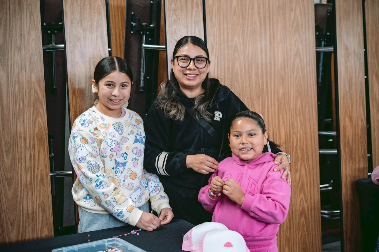 A woman with glasses and two young girls standing together indoors, with folded chairs and wooden walls in the background.