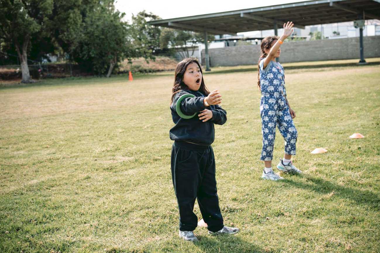 Two children playing football on a grassy field during daytime, with trees and a structure in the background.