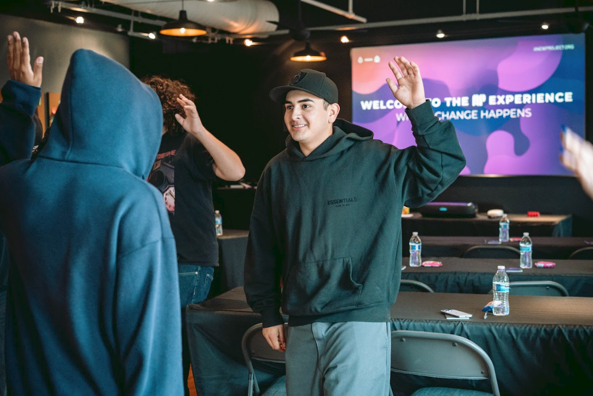 Young man in black hoodie and cap giving a high five to a person in a blue hoodie during an indoor event with tables, water bottles, and a large screen in the background