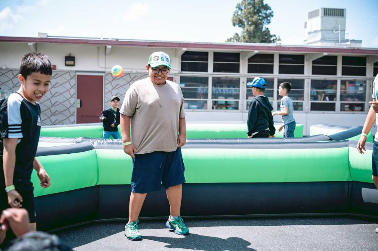 Children playing inside a green and black inflatable bounce house outdoors on a schoolyard or playground, some smiling and watching a colorful ball.