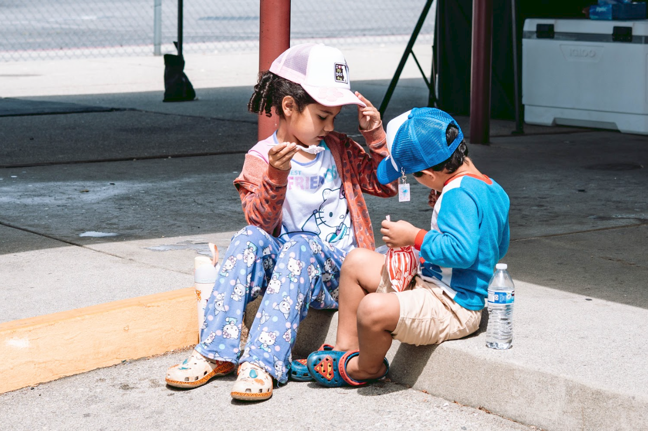 A young girl and boy sit on the sidewalk, sharing a snack. The girl has curly hair, wears a pink and white cap, a colorful jacket, and blue pajama pants with cartoon characters. The boy wears a blue cap backwards, a blue jacket, and tan shorts. They 