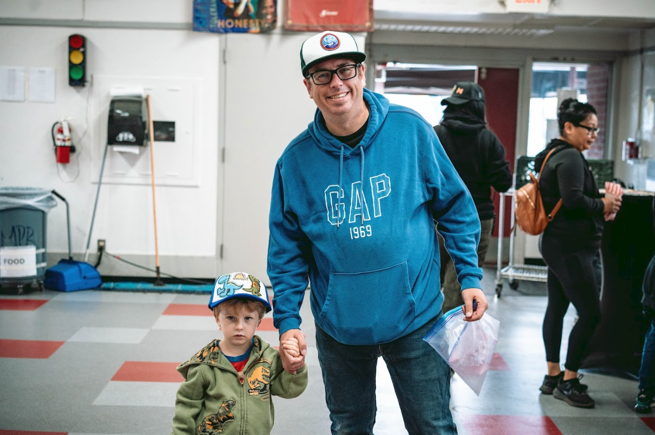 A man and a young boy holding hands inside a store or warehouse. The man is wearing a blue hoodie, black glasses, and a white and green cap, smiling at the camera. The boy is wearing a green jacket with dinosaur patches and a baseball cap with a cart