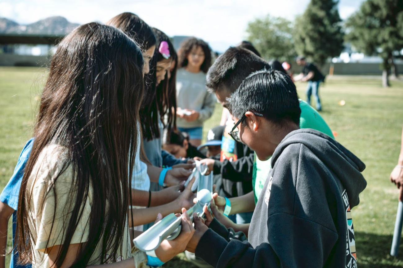 A group of children and teenagers gathered outdoors on a sunny day, some holding sheets of paper or posters, in a park or open field.
