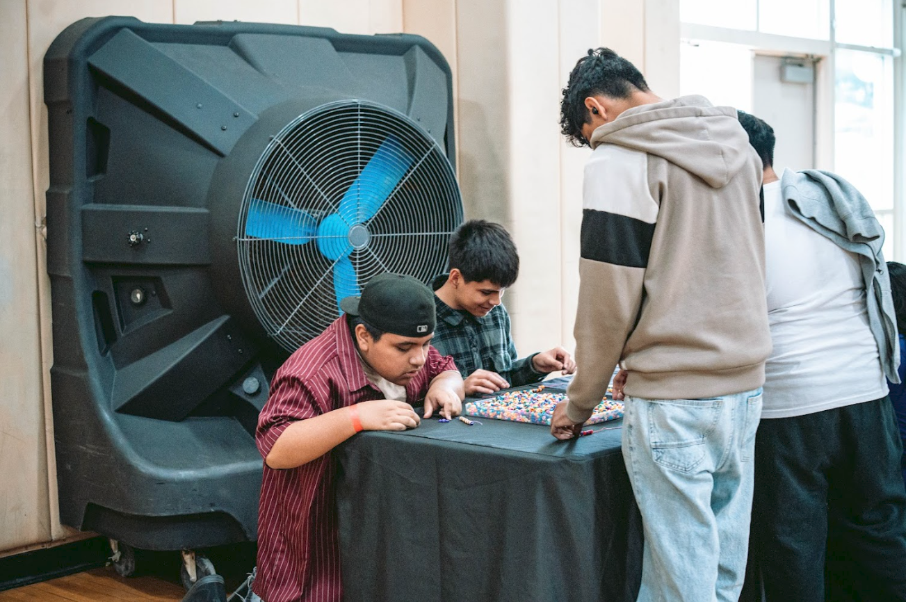 Four boys gathered around a table playing a bead maze game, indoors near a large fan and window.