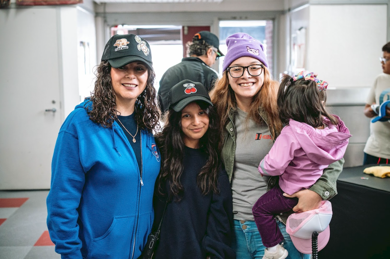 Group of four smiling females in casual clothing, standing indoors, with two women holding children, black and gray hats, colorful accessories, and a background with other people and objects.