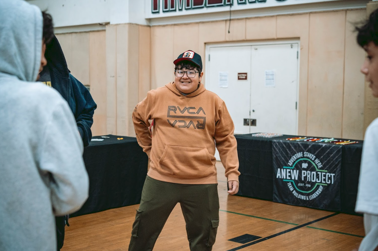 A smiling young man wearing glasses, a brown hoodie, and a black and red cap stands in a room with other people, with tables displaying items in the background.