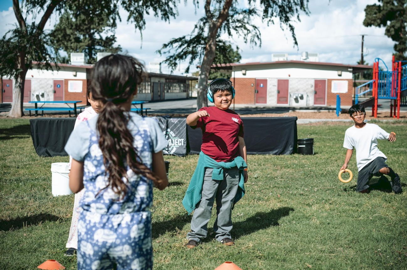 Children playing with ring toss game outdoors on a grassy field with school buildings and playground in the background.