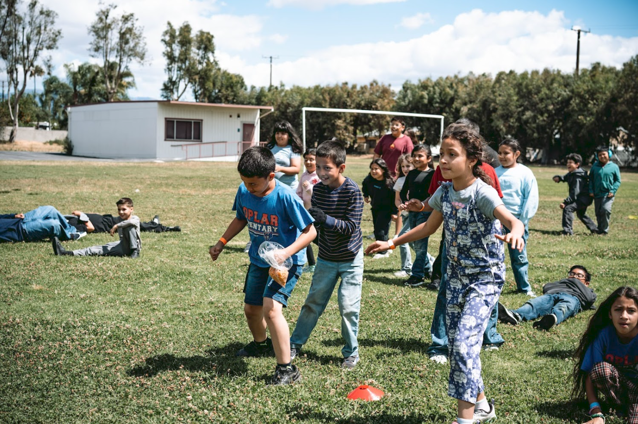 Children and adults playing outdoors on a grassy field during daytime with a small building and trees in the background.