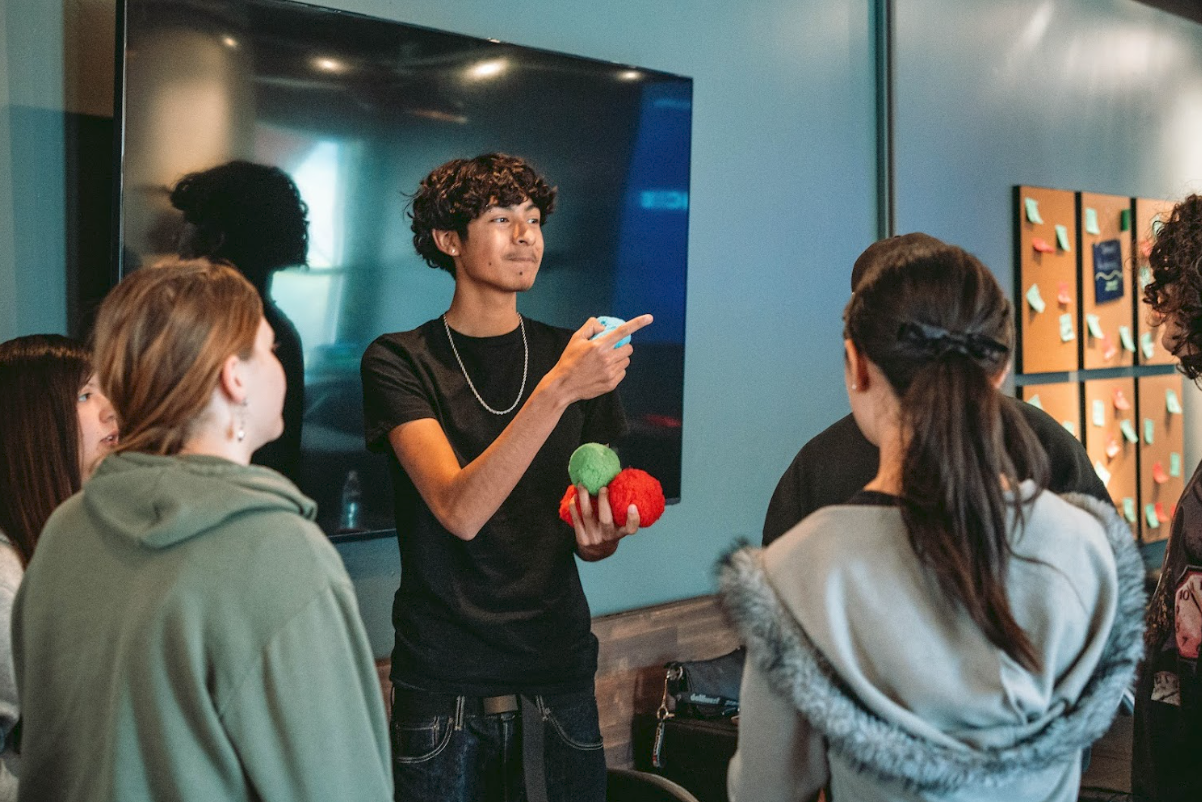 A young man with curly hair and a black shirt is holding colorful balls in one hand, pointing with the other, and appears to be demonstrating or explaining something to a group of people gathered around him in a room with a large monitor and bulletin