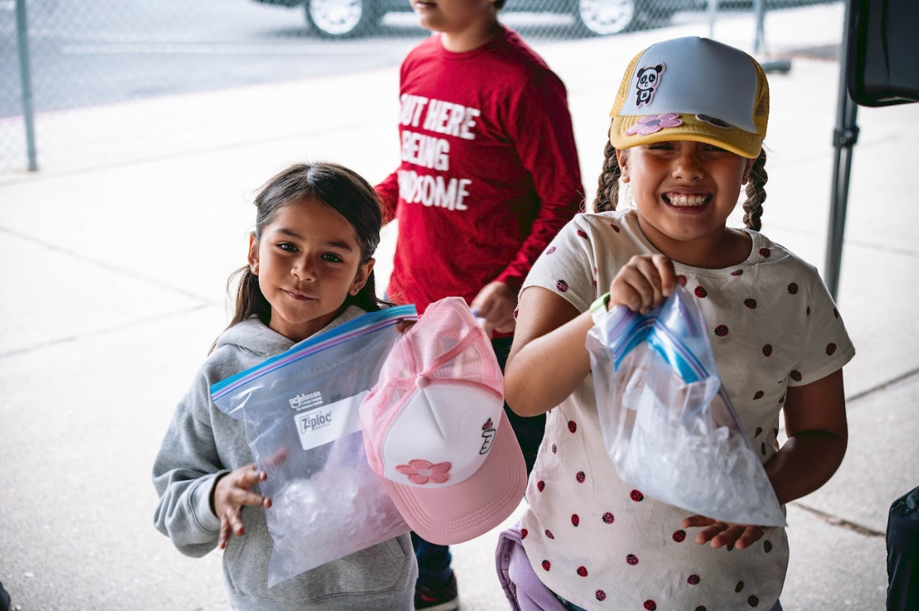 Three children standing outside near a chain-link fence, each holding a plastic bag with items inside, and one is wearing a pink cap.