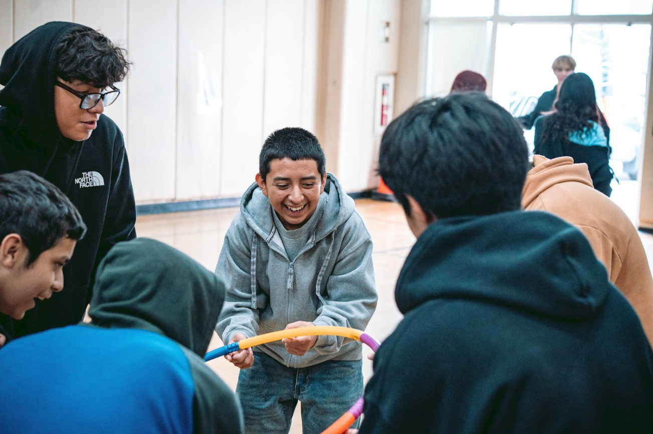 A group of young people in casual clothing, gathered around and engaging with each other in a circle, playing with colorful hula hoops indoors.