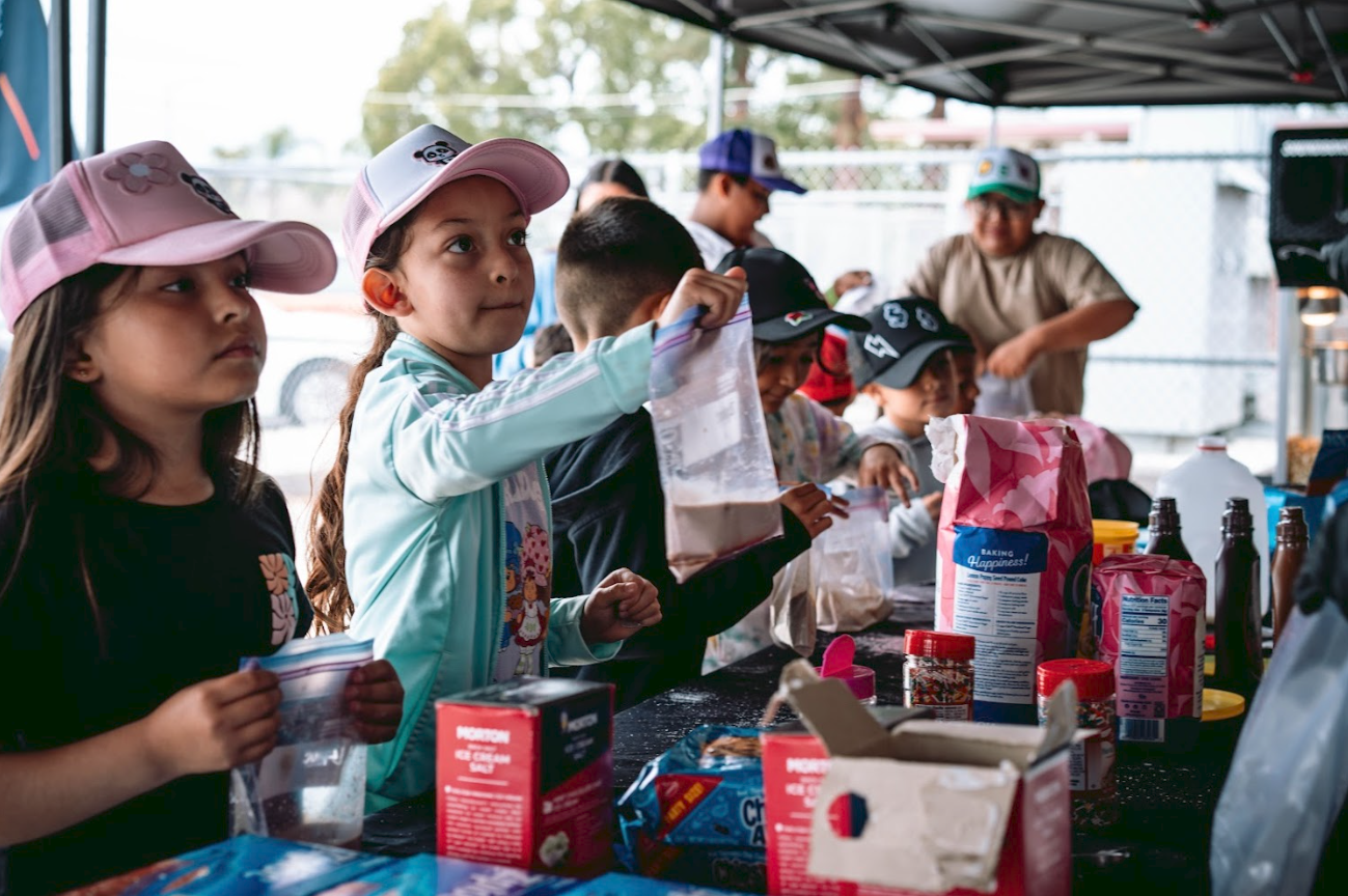 Group of children participating in a baking activity under a tent, with baking ingredients and supplies on the table.