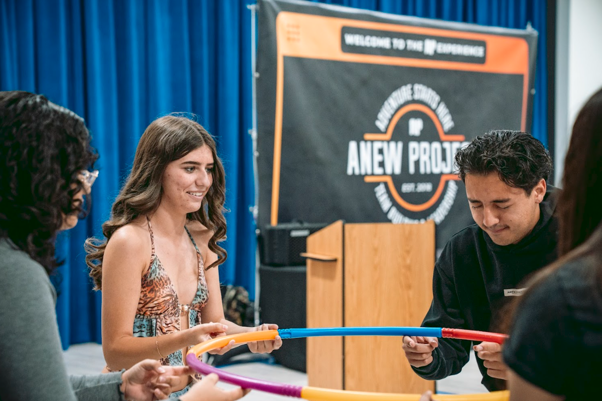 Group of young people participating in a team-building activity with colorful hula hoops at an indoor event with a black and orange sign in the background.