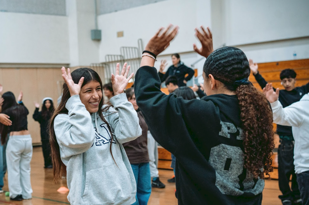 Two girls in a gymnasium playing a high-five game with children in the background.