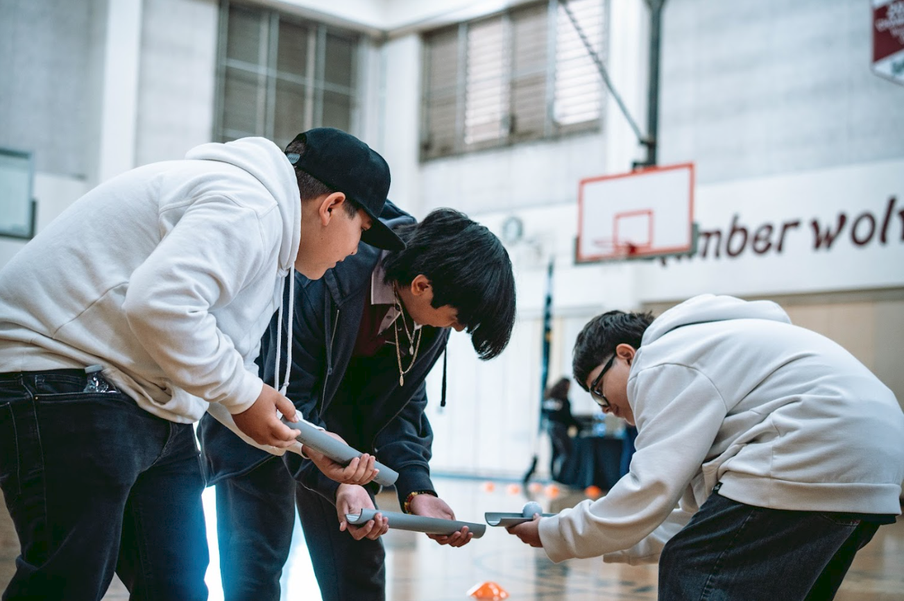 Three young men are in a gym, holding and examining flat, round objects, possibly awards or medals, during an indoor sports event.