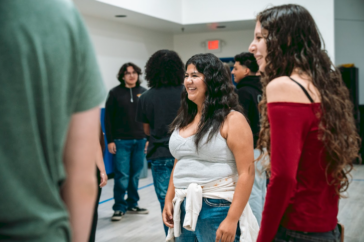A group of diverse young people gathered indoors, smiling and engaging with each other, with a woman in a gray tank top and a woman in a red off-shoulder top in the foreground.