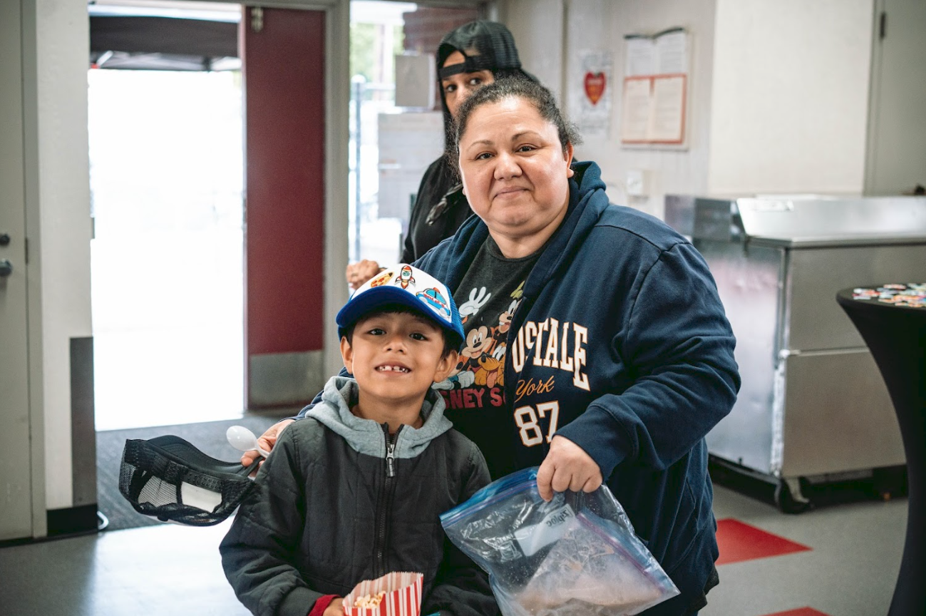 A woman and a young boy standing inside a room, with both smiling. The woman is holding a plastic bag, and the boy is holding a popcorn container. Behind them, a woman is partially visible, and there are bulletin boards and a table with items on it.