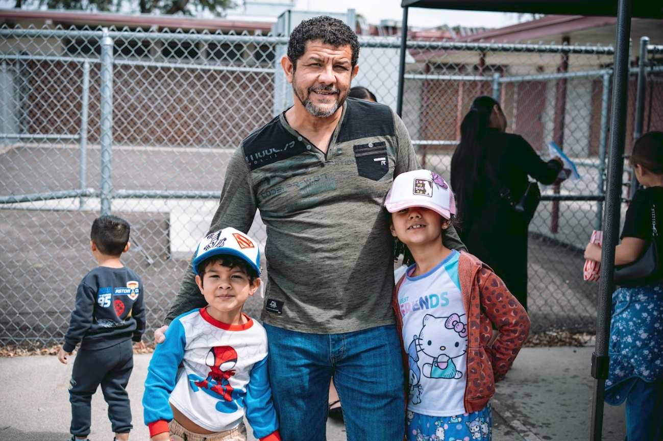 A man with two children at an outdoor playground or park, with a chain-link fence in the background. The man is smiling and wearing a gray and black shirt, and the children are wearing colorful shirts and hats.