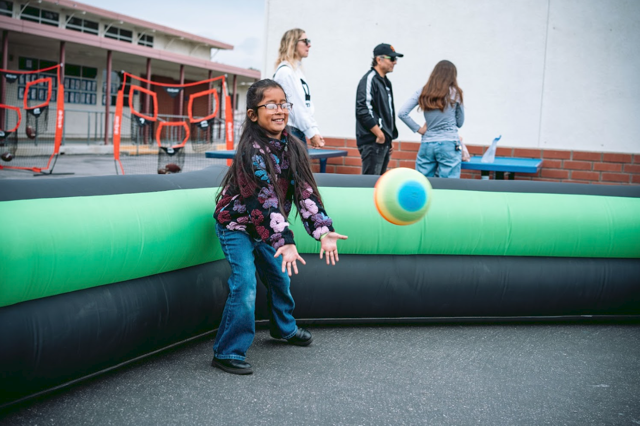 A young girl playing with a colorful ball in a bounce house at an outdoor event, with other children and adults in the background near a brick wall and a fun activity area with red ring toss game.