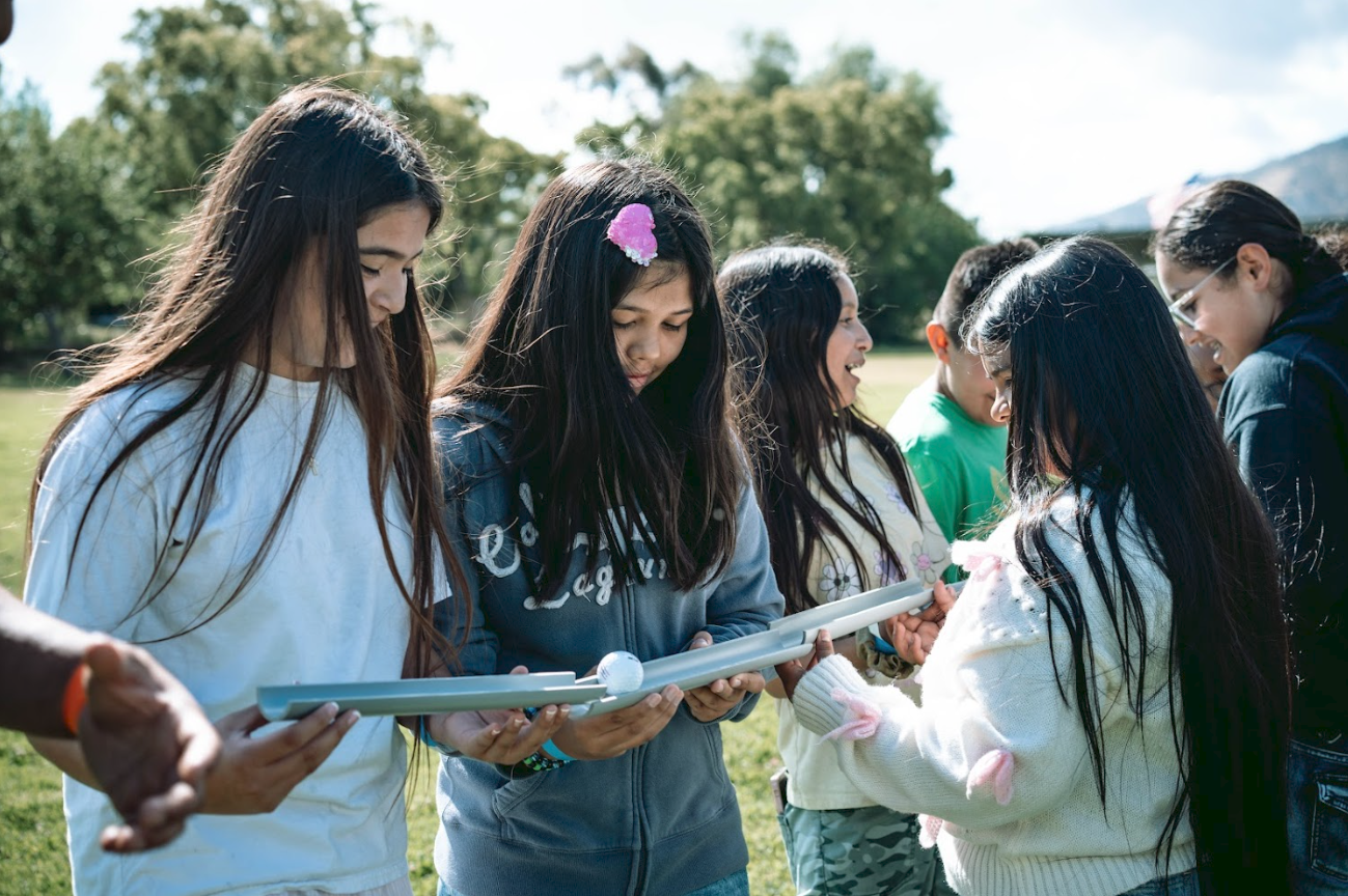 A group of diverse young girls standing outdoors in a park, engaged in observing a small object, possibly a toy or learning tool, that one girl is holding. They are smiling and seem interested in the activity, with trees and mountains in the backgrou