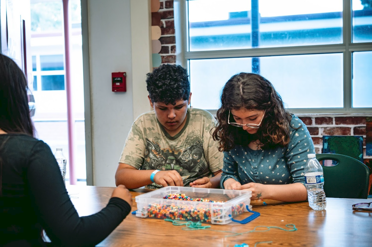Two children and an adult sitting at a table, working on a craft project with colorful beads and strings, in a room with brick and window walls.