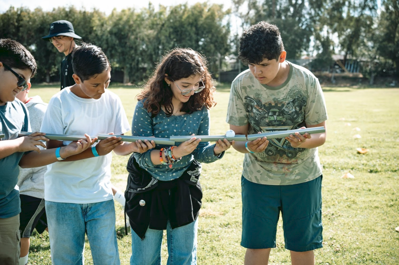 Group of children outdoors holding a long blue tube, with a girl smiling and a boy looking at the tube.
